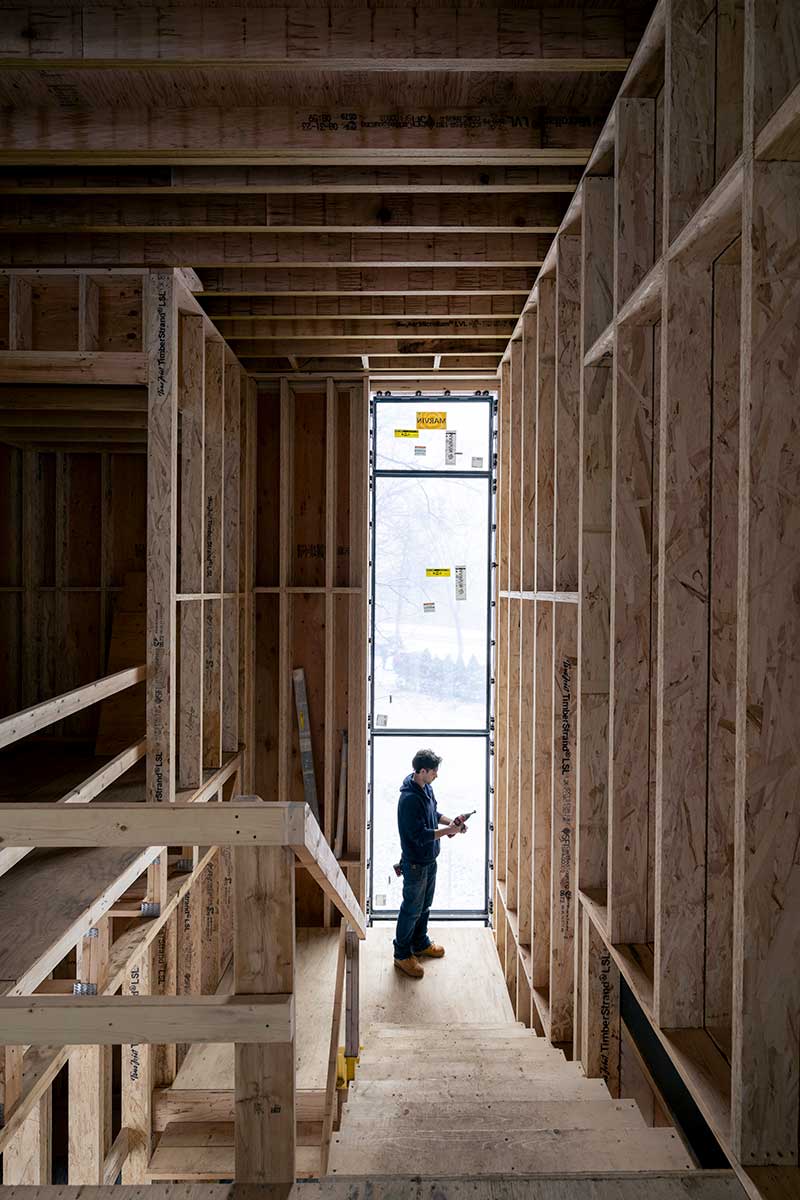 A man stands next to a floor-to-ceiling Marvin Modern window at the bottom of a staircase in an under construction home.